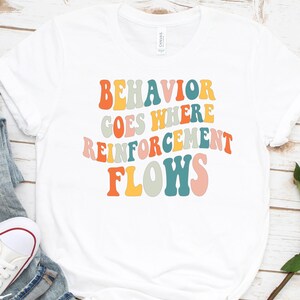 May include: White t-shirt with the text "BEHAVIOR GOES WHERE REINFORCEMENT FLOWS" in colorful retro font.