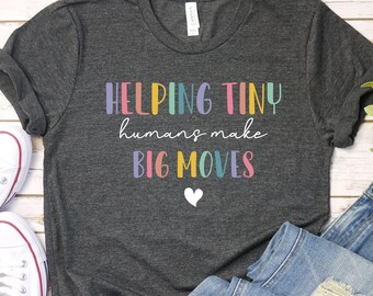 May include: A dark grey t-shirt with the text "HELPING TINY humans make BIG MOVES" in rainbow colours. A small white heart is below the text.