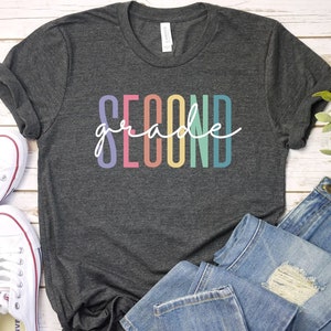May include: A dark gray t-shirt with the text "second grade" in rainbow colors. The shirt is laid out on a white wooden surface with a pair of white sneakers and blue jeans.