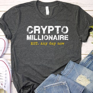May include: A dark gray t-shirt with the text "CRYPTO MILLIONAIRE EST. Any day now" in white and yellow lettering. The shirt is laid out on a white wooden surface with a pair of white sneakers, a pair of blue jeans, and a small green plant.