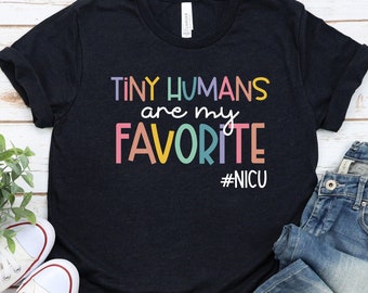May include: Black t-shirt with the text "Tiny Humans are my Favorite #NICU" in rainbow colors. The shirt is folded and laid on a wooden surface with a pair of white sneakers and blue jeans.