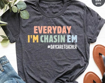 Puede incluir: Camiseta gris oscuro jaspeada con el texto "EVERYDAY I'M CHASIN' EM #DAYCARETEACHER" en colores arcoíris.