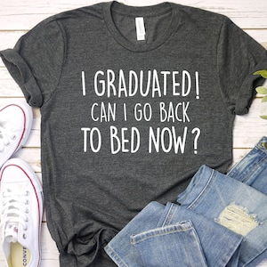May include: A dark gray t-shirt with white text that reads "I graduated! Can I go back to bed now?" The shirt is laid flat on a white wooden surface with a pair of white sneakers and blue jeans.