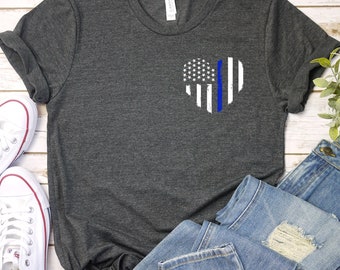 Könnte beinhalten: Ein dunkelgraues T-Shirt mit einem weißen Herzdesign, das eine amerikanische Flagge mit einem blauen Streifen in der Mitte zeigt. Das Shirt liegt auf einer weißen Holzoberfläche mit einem Paar weißen Turnschuhen und einer blauen Jeans.