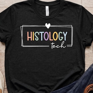 May include: Black t-shirt with the words "HISTOLOGY tech" in a rectangular box with a heart above it. The text is in pastel colors. The shirt is paired with blue jeans and sneakers.