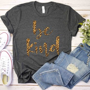 May include: A gray t-shirt with the words "be kind" printed in a leopard print design. The shirt is laid out on a white wooden surface with a pair of white sneakers and a pair of blue jeans.