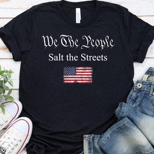 May include: A navy blue t-shirt with the text "We The People Salt the Streets" and a distressed American flag graphic. The shirt is paired with blue jeans and white sneakers.
