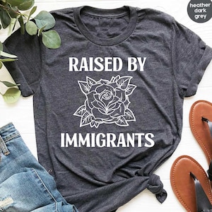 May include: Dark heather grey t-shirt with the text "RAISED BY IMMIGRANTS" in white, featuring a rose graphic. The shirt is paired with blue jeans and brown sandals. The shirt is made of a soft, comfortable material.