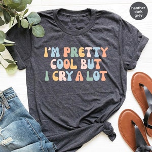 May include: Dark heather grey t-shirt with the text "I'M PRETTY COOL BUT I CRY A LOT" in colorful retro font. The shirt is displayed with blue jeans and brown sandals.