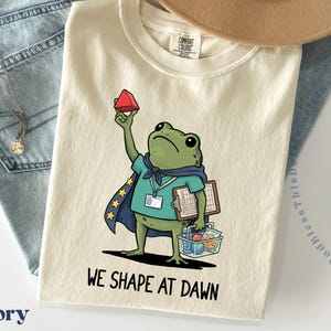 May include: An ivory t-shirt featuring a graphic of a green frog wearing a blue cape with yellow stars, a teal shirt, and a lanyard. The frog holds a red triangular prism, a clipboard, and a basket containing various shapes. Text reads WE SHAPE AT DAWN.