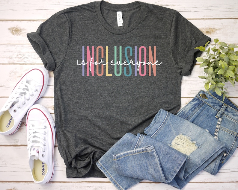 May include: A dark grey t-shirt with the text "Inclusion is for everyone" in rainbow colors. The shirt is laid out on a white wooden surface with a pair of white sneakers and blue jeans.
