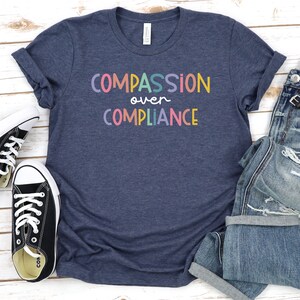 Pode incluir: Uma camiseta azul marinho com o texto "COMPASSION over COMPLIANCE" em cores do arco-&iacute;ris. A camiseta est&aacute; colocada sobre uma superf&iacute;cie de madeira branca com um par de sapatos de lona pretos e um par de cal&ccedil;as de ganga azuis.