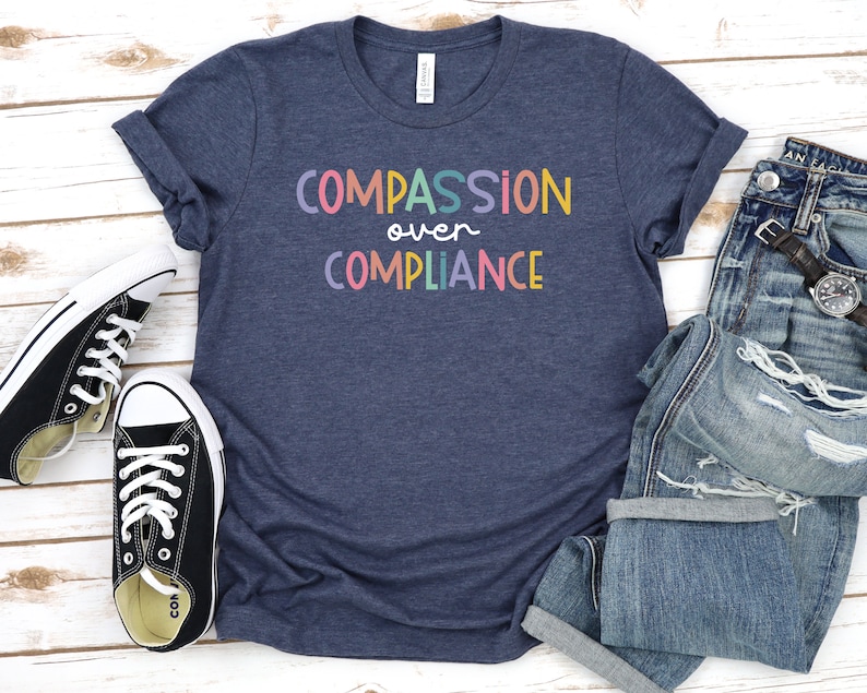 Pode incluir: Uma camiseta azul marinho com o texto "COMPASSION over COMPLIANCE" em cores do arco-&iacute;ris. A camiseta est&aacute; colocada sobre uma superf&iacute;cie de madeira branca com um par de sapatos de lona pretos e um par de cal&ccedil;as de ganga azuis.