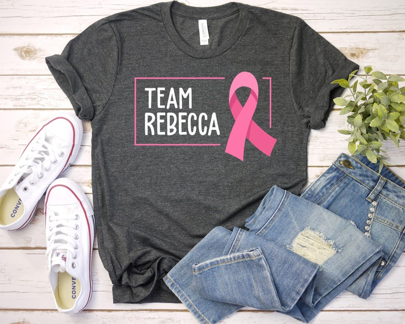 Pode incluir: Camiseta cinza com o texto "TEAM REBECCA" em uma caixa rosa e uma fita rosa. A camiseta est&aacute; colocada em uma superf&iacute;cie de madeira branca com um par de t&ecirc;nis brancos e uma cal&ccedil;a jeans azul.