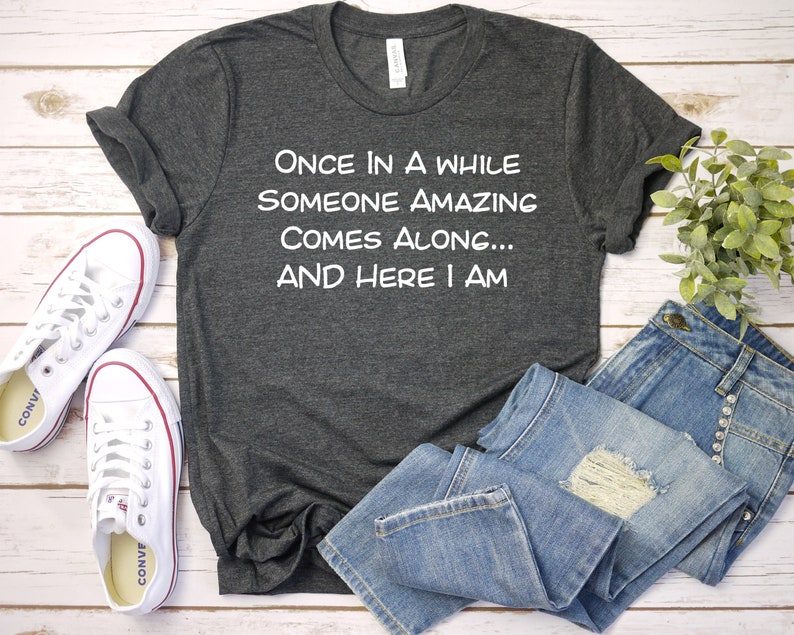 Peut inclure: Un t-shirt gris fonc&eacute; avec un texte blanc qui dit "Once in a while someone amazing comes along... And here I am". Le t-shirt est pos&eacute; sur une surface en bois blanc avec une paire de baskets blanches et un jean bleu.