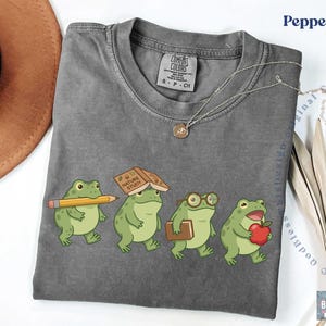 K&ouml;nnte beinhalten: Ein anthrazitfarbenes Kurzarm-T-Shirt mit einer Grafik von vier gr&uuml;nen Fr&ouml;schen. Ein Frosch h&auml;lt einen gelben Bleistift, einer hat ein Buch mit dem Titel Nature Study auf dem Kopf, einer tr&auml;gt eine Brille und ein Buch, und einer h&auml;lt einen roten Apfel.