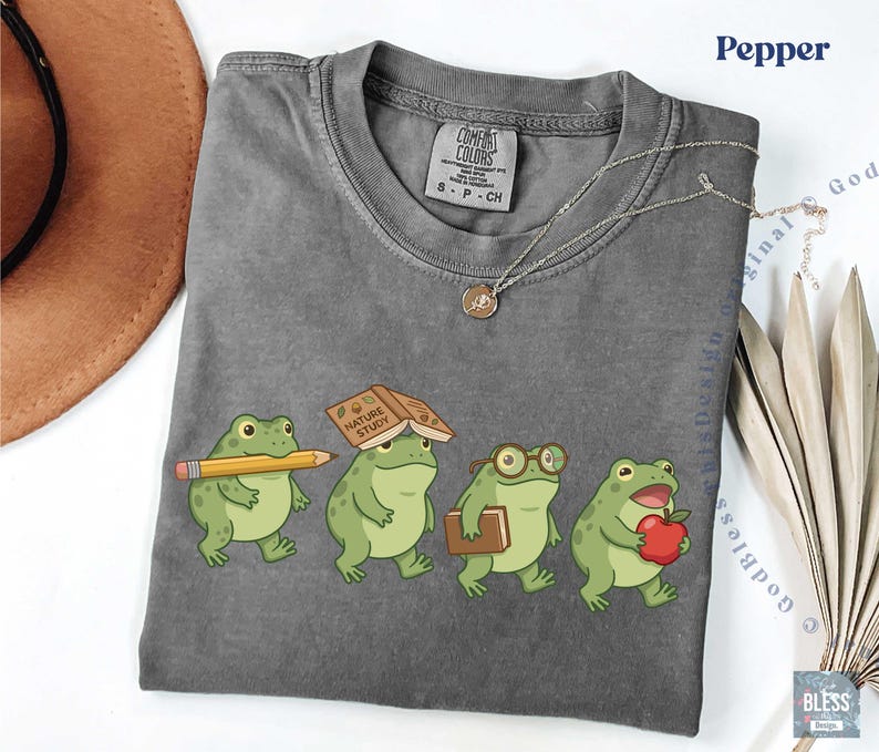K&ouml;nnte beinhalten: Ein anthrazitfarbenes Kurzarm-T-Shirt mit einer Grafik von vier gr&uuml;nen Fr&ouml;schen. Ein Frosch h&auml;lt einen gelben Bleistift, einer hat ein Buch mit dem Titel Nature Study auf dem Kopf, einer tr&auml;gt eine Brille und ein Buch, und einer h&auml;lt einen roten Apfel.