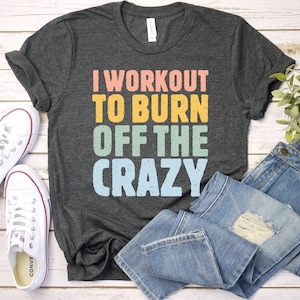 May include: A dark gray t-shirt with the text "I workout to burn off the crazy" in colorful, distressed lettering. The shirt is laid out on a white wooden surface with a pair of white sneakers and blue jeans.
