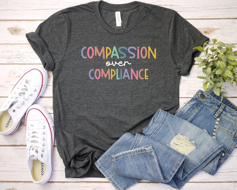 Pode incluir: Uma camiseta cinza escura com o texto "COMPASSION over COMPLIANCE" em letras coloridas. A camiseta est&aacute; colocada sobre uma superf&iacute;cie de madeira branca com um par de t&ecirc;nis brancos e uma cal&ccedil;a jeans azul.