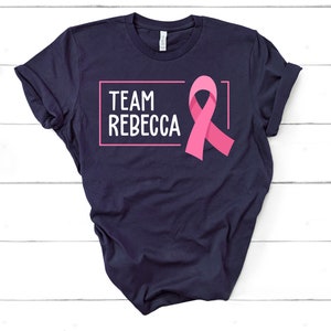 Pode incluir: Camiseta azul marinho com uma fita rosa e o texto "TEAM REBECCA" em uma caixa branca.