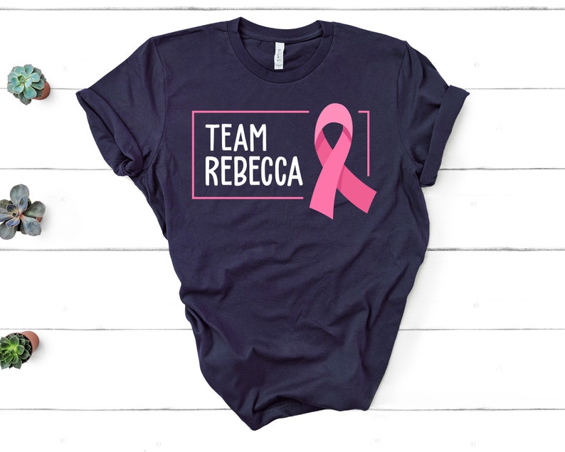 Pode incluir: Camiseta azul marinho com uma fita rosa e o texto "TEAM REBECCA" em uma caixa branca.