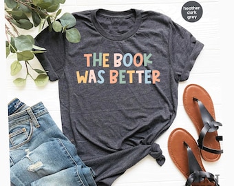 May include: Dark gray heather t-shirt with the text "THE BOOK WAS BETTER" in colorful letters. The shirt is paired with blue jeans and brown sandals, suggesting a casual, everyday style. The design is suitable for book lovers.
