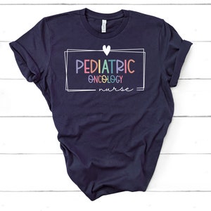 Peut inclure: Un t-shirt bleu marine avec un c&oelig;ur blanc et un cadre blanc autour du texte "PEDIATRIC ONCOLOGY nurse". Le texte est en couleurs arc-en-ciel.