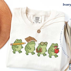K&ouml;nnte beinhalten: Ein elfenbeinfarbenes T-Shirt mit einer Grafik von vier gr&uuml;nen Fr&ouml;schen. Ein Frosch h&auml;lt einen gelben Bleistift, einer hat ein Buch mit dem Titel Nature Study auf dem Kopf, einer tr&auml;gt eine Brille und ein Buch, und einer h&auml;lt einen roten Apfel. Comfort Colors Etikett.