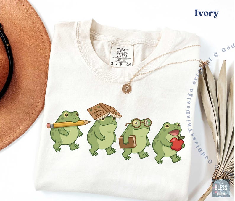 K&ouml;nnte beinhalten: Ein elfenbeinfarbenes T-Shirt mit einer Grafik von vier gr&uuml;nen Fr&ouml;schen. Ein Frosch h&auml;lt einen gelben Bleistift, einer hat ein Buch mit dem Titel Nature Study auf dem Kopf, einer tr&auml;gt eine Brille und ein Buch, und einer h&auml;lt einen roten Apfel. Comfort Colors Etikett.