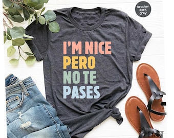 May include: Heather dark grey t-shirt with the text "I'M NICE PERO NO TE PASES" in colorful block letters. The shirt is displayed with blue jeans and brown sandals.