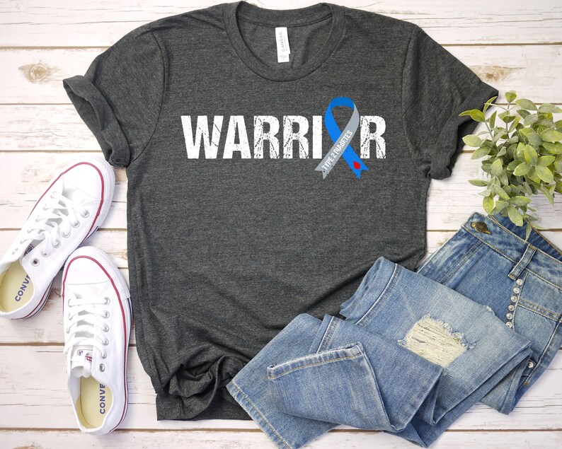 K&ouml;nnte beinhalten: Ein dunkelgraues T-Shirt mit dem Wort "WARRIOR" in wei&szlig; und einem Used-Look-Effekt bedruckt. Auf der rechten Seite des Shirts ist ein blaues Band mit dem Text "TYPE 1 DIABETES" aufgedruckt.
