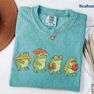 K&ouml;nnte beinhalten: Ein meergr&uuml;nes T-Shirt mit Rundhalsausschnitt und einer Grafik von vier Cartoon-Fr&ouml;schen. Ein Frosch h&auml;lt einen gelben Bleistift, einer hat ein Buch mit dem Titel Nature Study auf dem Kopf, einer tr&auml;gt eine Brille und einer h&auml;lt einen roten Apfel.
