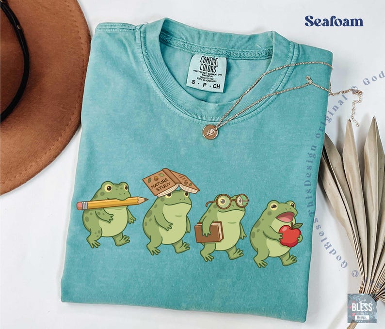 K&ouml;nnte beinhalten: Ein meergr&uuml;nes T-Shirt mit Rundhalsausschnitt und einer Grafik von vier Cartoon-Fr&ouml;schen. Ein Frosch h&auml;lt einen gelben Bleistift, einer hat ein Buch mit dem Titel Nature Study auf dem Kopf, einer tr&auml;gt eine Brille und einer h&auml;lt einen roten Apfel.