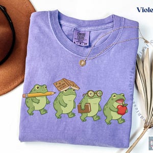 K&ouml;nnte beinhalten: Ein violettes Kurzarm-T-Shirt mit einer Grafik von vier gr&uuml;nen Fr&ouml;schen. Ein Frosch h&auml;lt einen gelben Bleistift, einer hat ein Buch mit dem Titel Nature Study auf dem Kopf, einer tr&auml;gt eine Brille und ein Buch, und einer h&auml;lt einen roten Apfel.