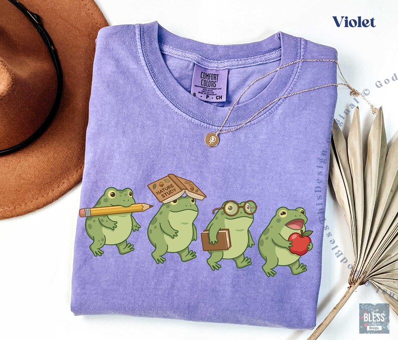 K&ouml;nnte beinhalten: Ein violettes Kurzarm-T-Shirt mit einer Grafik von vier gr&uuml;nen Fr&ouml;schen. Ein Frosch h&auml;lt einen gelben Bleistift, einer hat ein Buch mit dem Titel Nature Study auf dem Kopf, einer tr&auml;gt eine Brille und ein Buch, und einer h&auml;lt einen roten Apfel.