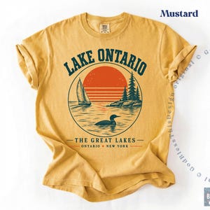 May include: A mustard yellow short-sleeve t-shirt featuring a graphic of Lake Ontario. The design includes a sailboat, a loon, pine trees, and a red sun within a circle. Text reads LAKE ONTARIO, THE GREAT LAKES, and ONTARIO NEW YORK.