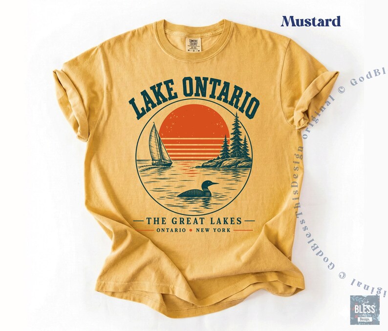 May include: A mustard yellow short-sleeve t-shirt featuring a graphic of Lake Ontario. The design includes a sailboat, a loon, pine trees, and a red sun within a circle. Text reads LAKE ONTARIO, THE GREAT LAKES, and ONTARIO NEW YORK.