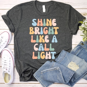 May include: A dark gray t-shirt with the text "Shine Bright Like A Call Light" in colorful, retro-style lettering.