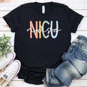 May include: A black t-shirt with the text "NICU nurse" in rainbow colors. The shirt is laid flat on a white wooden surface with a pair of white sneakers and blue jeans.