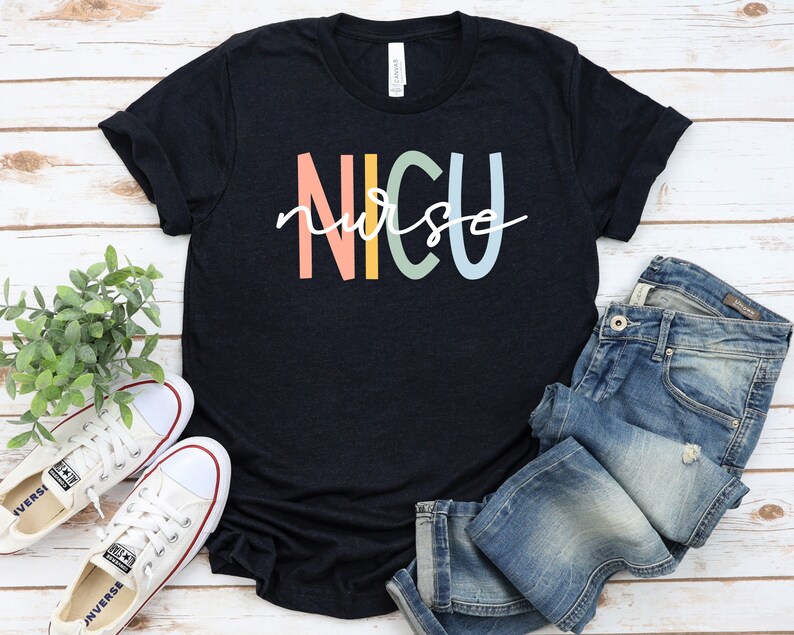 May include: A black t-shirt with the text "NICU nurse" in rainbow colors. The shirt is laid flat on a white wooden surface with a pair of white sneakers and blue jeans.