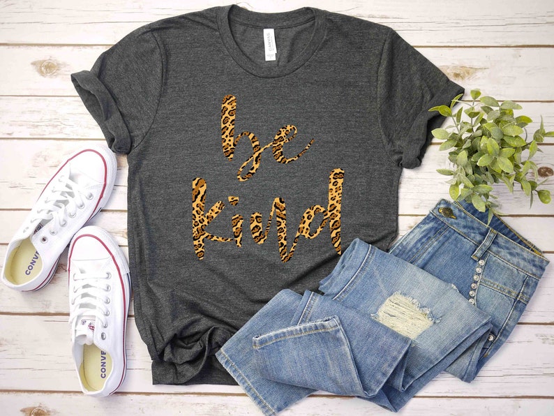 May include: A gray t-shirt with the words "be kind" printed in a leopard print design. The shirt is laid out on a white wooden surface with a pair of white sneakers and a pair of blue jeans.