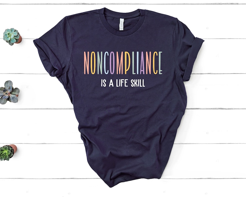 K&ouml;nnte beinhalten: Marineblaues T-Shirt mit dem Text "NONCOMPLIANCE IS A LIFE SKILL" in bunten Pastellbuchstaben. Das Shirt liegt auf einer wei&szlig;en Holzoberfl&auml;che, im Hintergrund sind kleine Sukkulenten in T&ouml;pfen zu sehen.
