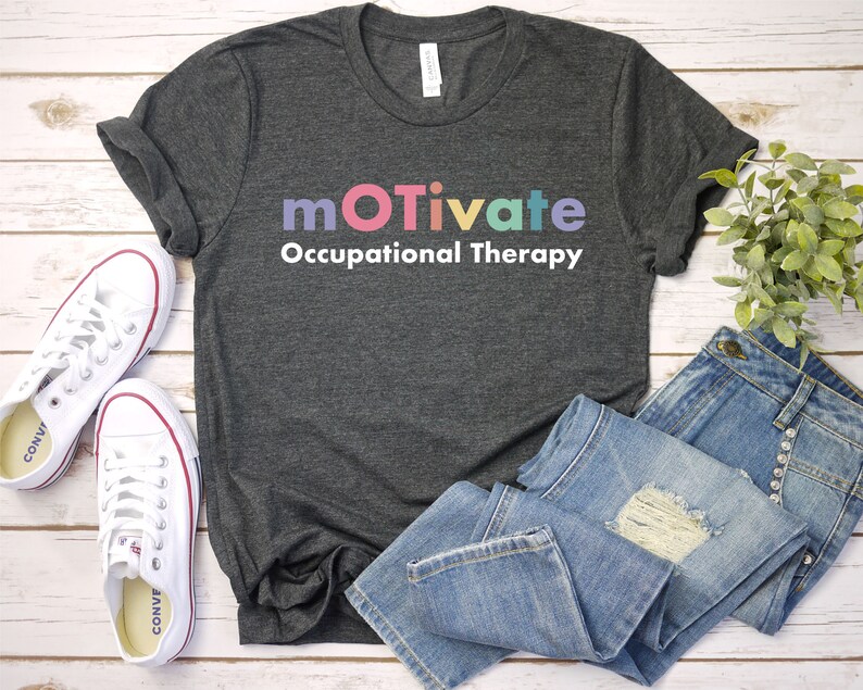 May include: A dark grey t-shirt with the text "motivate Occupational Therapy" in rainbow colours. The shirt is laid out on a white wooden surface with a pair of white trainers and blue jeans.