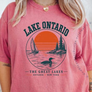 May include: A coral-colored short-sleeve t-shirt featuring a circular graphic of Lake Ontario with a sailboat, a loon, pine trees, and an orange sun. The text reads LAKE ONTARIO, THE GREAT LAKES, and ONTARIO NEW YORK in dark green lettering.
