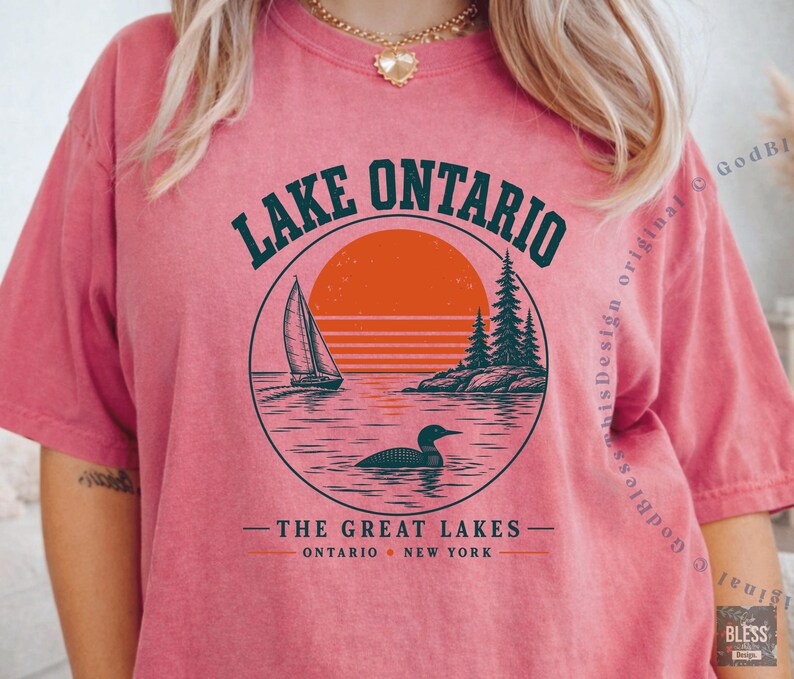 May include: A coral-colored short-sleeve t-shirt featuring a circular graphic of Lake Ontario with a sailboat, a loon, pine trees, and an orange sun. The text reads LAKE ONTARIO, THE GREAT LAKES, and ONTARIO NEW YORK in dark green lettering.