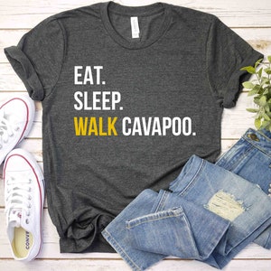 May include: A dark gray t-shirt with the text "Eat. Sleep. Walk Cavapoo." printed in white on the front. The shirt is laid flat on a white wooden surface with a pair of white sneakers and blue jeans.