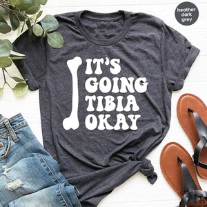May include: A heather dark grey t-shirt with a white bone graphic and the text "It's Going Tibia Okay" in a retro font.
