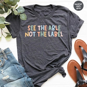 Pode incluir: Camiseta cinza escuro heather com o texto "See the able not the label" em cores do arco-íris.