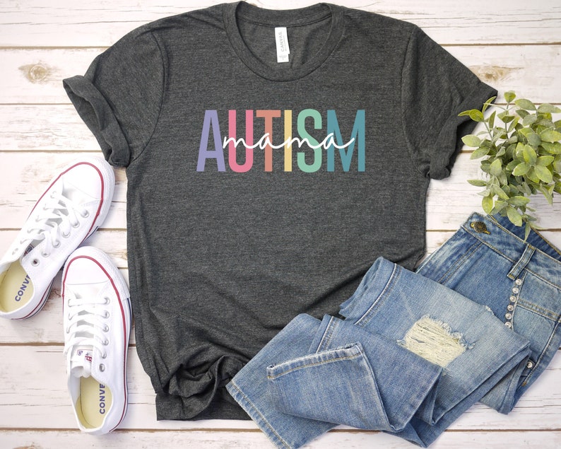 May include: A dark gray t-shirt with the text "AUTISM mama" in rainbow colors. The shirt is laid out on a white wooden surface with a pair of white sneakers and blue jeans.