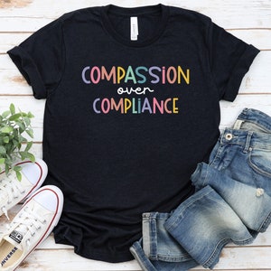 Pode incluir: Uma camiseta preta com o texto "COMPASSION over COMPLIANCE" em cores do arco-&iacute;ris. A camiseta est&aacute; colocada sobre uma superf&iacute;cie de madeira com um par de t&ecirc;nis brancos e uma cal&ccedil;a jeans azul.